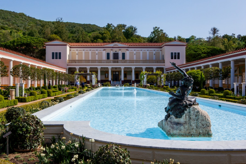 Bronze statue of a reclining male figure on a travertine slab sits in the middle of a shallow pool. Behind it is a garden in front of a covered walkway with four doric columns.